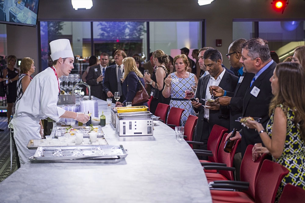 Guests stand at bar as chef plates meals