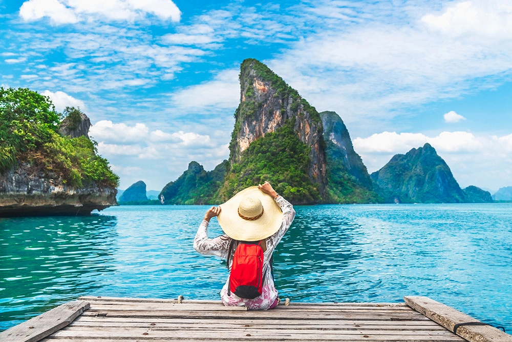 Woman with large straw hat and backpack sit on a dock looking at tropical scenery